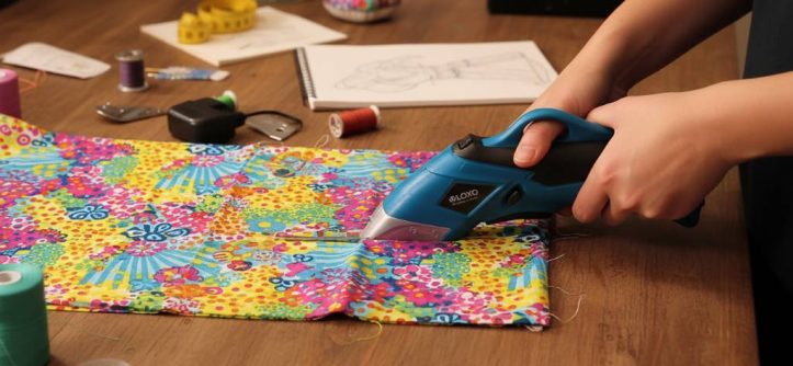 Person using cordless electric scissors to cut colorful fabric on a crafting table